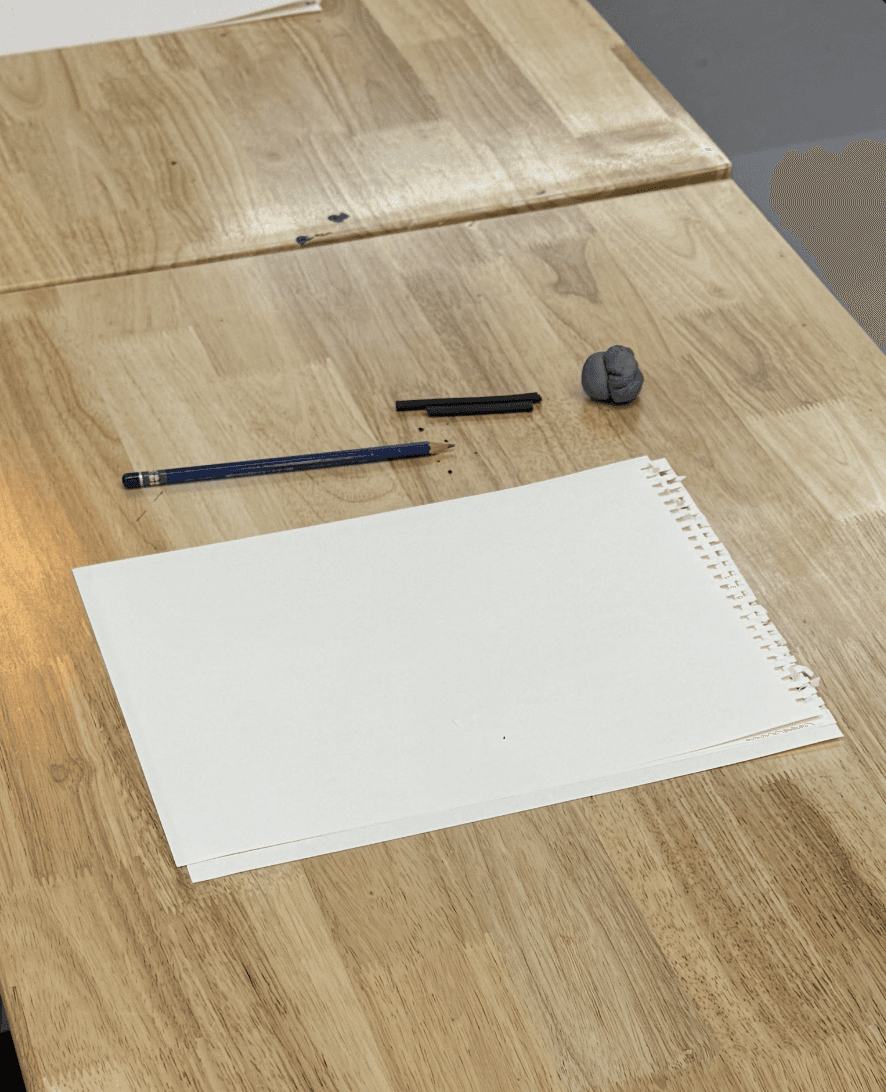 A close-up of a studio table set for class, with a blank sketchpad, pencil, charcoal, and a kneaded eraser laid out and ready